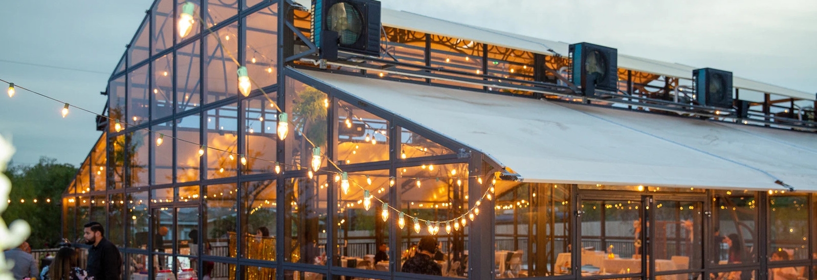 Elegant glass greenhouse venue adorned with string lights at dusk, showcasing interior warm lighting and exterior spot cooling units.